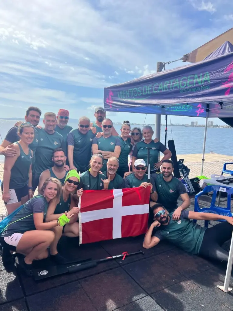 Tripulación de dragon boat del Club Náutico Santa Lucía con la bandera de Cartagena y una pala en el pantalán del Festival Jan Collins, con el Mar Menor al fondo en San Pedro del Pinatar