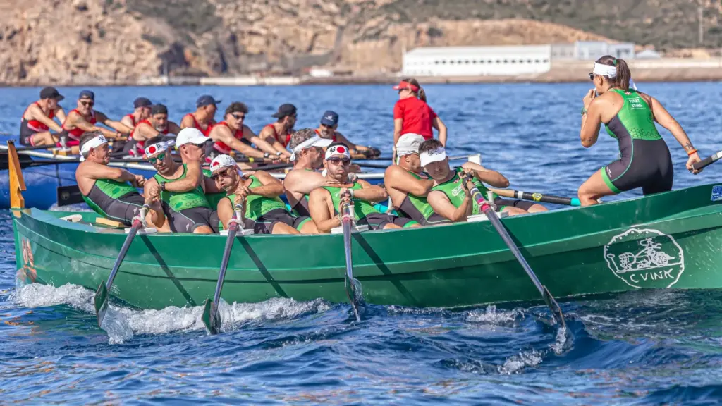 Tripulación masculina de remo de banco fijo con equipación verde y negra y patrona de pie, remando en un bote verde en el puerto de Cartagena durante la IX Travesía a Remo en Banco Fijo Puerto de Cartagena - Isla de las Palomas, seguida de cerca por otra embarcación de rojo