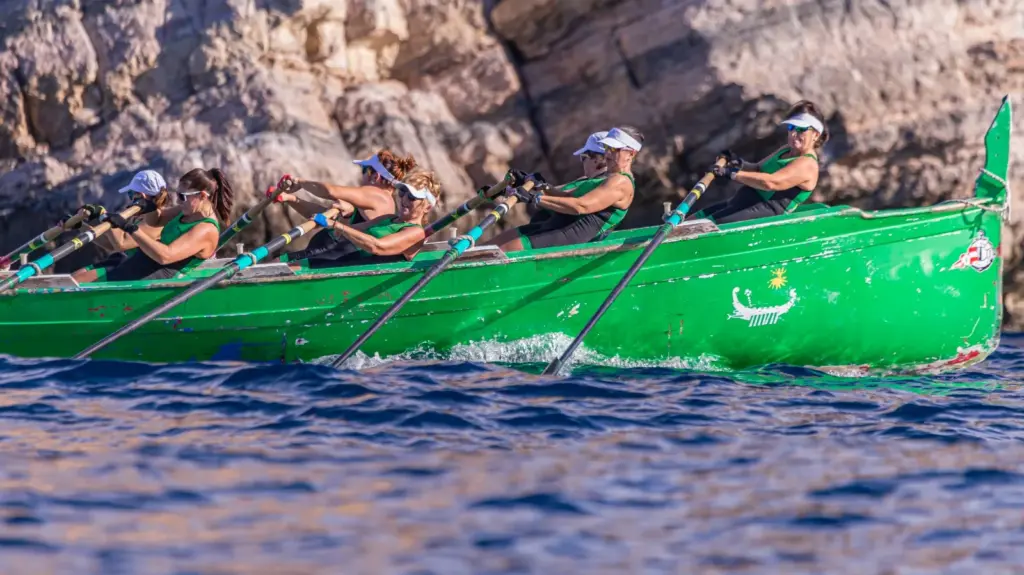 Tripulación femenina de remo de banco fijo con equipación verde remando en un bote verde junto a los acantilados rocosos de la costa de Cartagena durante la IX Travesía a Remo en Banco Fijo Puerto de Cartagena - Isla de las Palomas